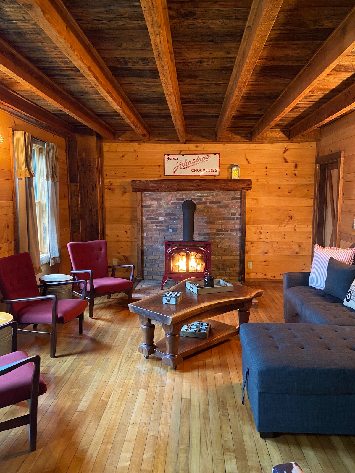 A cozy living area features a mix of seating, including red chairs, a dark sofa, and a wooden coffee table. A wood-burning stove with a visible flame is positioned against a rustic stone accent wall, enhancing the warm ambiance of the room.