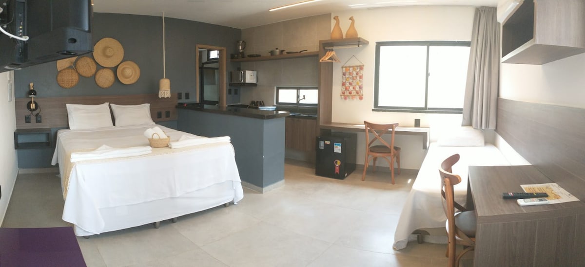 A modern studio apartment features two spacious beds with white linens and natural wood accents. A kitchenette is visible in the background with essential appliances. A window invites natural light, complemented by a workspace with a wooden desk and a chair nearby.