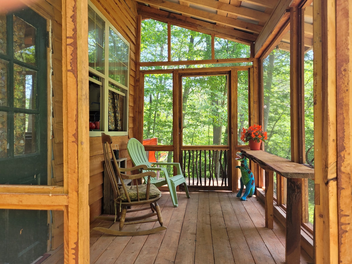 A welcoming front porch features wooden flooring and walls made of large windows, allowing natural light to fill the space. Two chairs are positioned for relaxation, accompanied by potted flowers that add a touch of color. A wooden railing enhances the view of the surrounding forest.