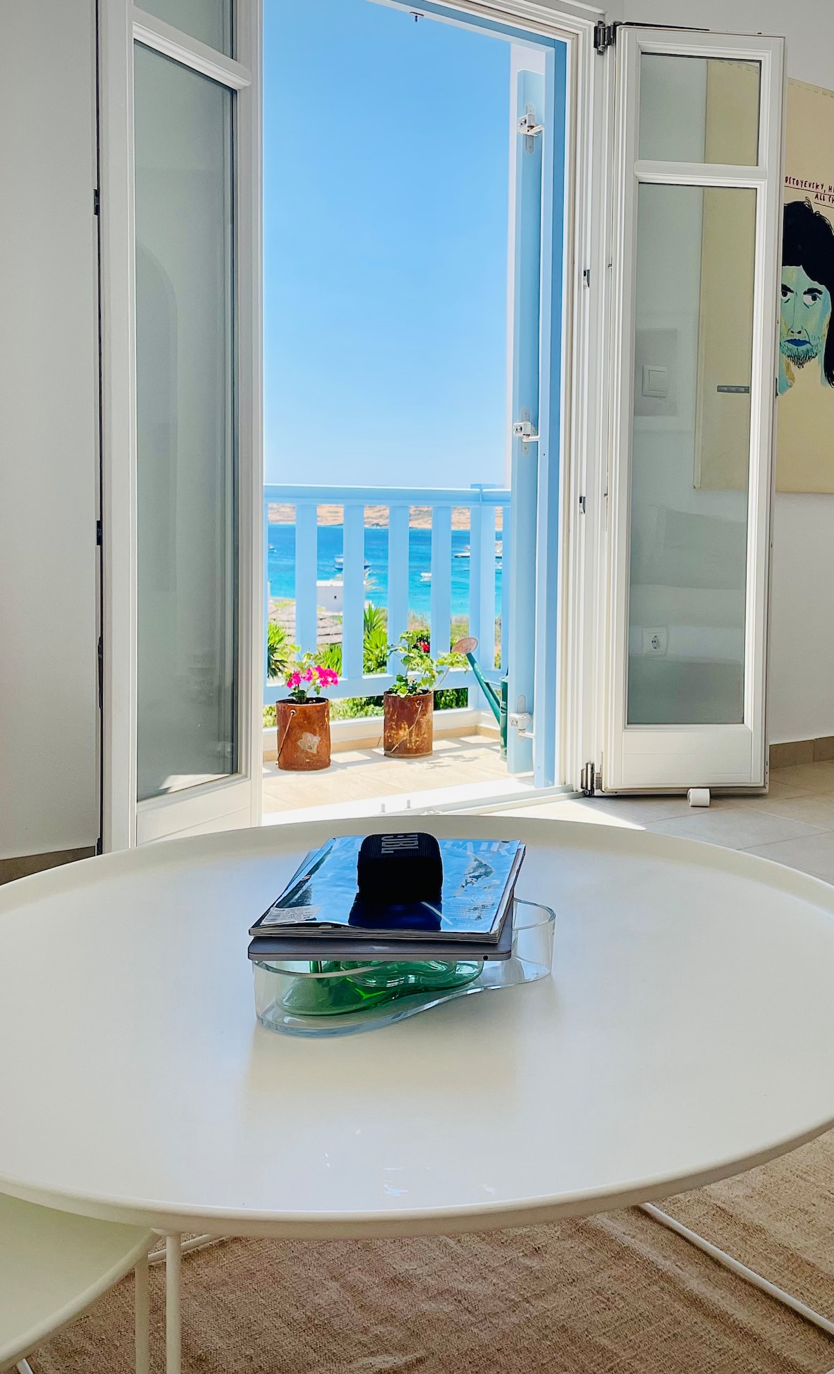 A bright living area features a modern white table centered in the foreground. Open doors reveal a stunning view of the sea, with blue waters and colorful flowers visible on the balcony. A sleek container rests on the table, adding a touch of functionality.