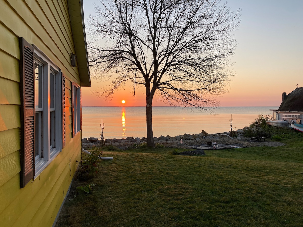 A yellow cottage stands beside tranquil waters at sunset, with the sun reflected on the calm lake surface. A bare tree is positioned in the foreground, and a grassy yard leads to a rocky shoreline, creating a serene lakeside ambiance.