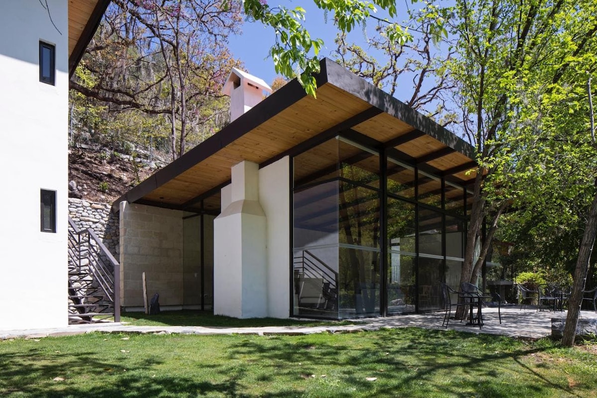 A modern structure with large glass windows is surrounded by greenery. A stone pathway leads to the entrance, while a spacious outdoor area features a manicured lawn. The roof extends outward, creating a shaded space for relaxation.