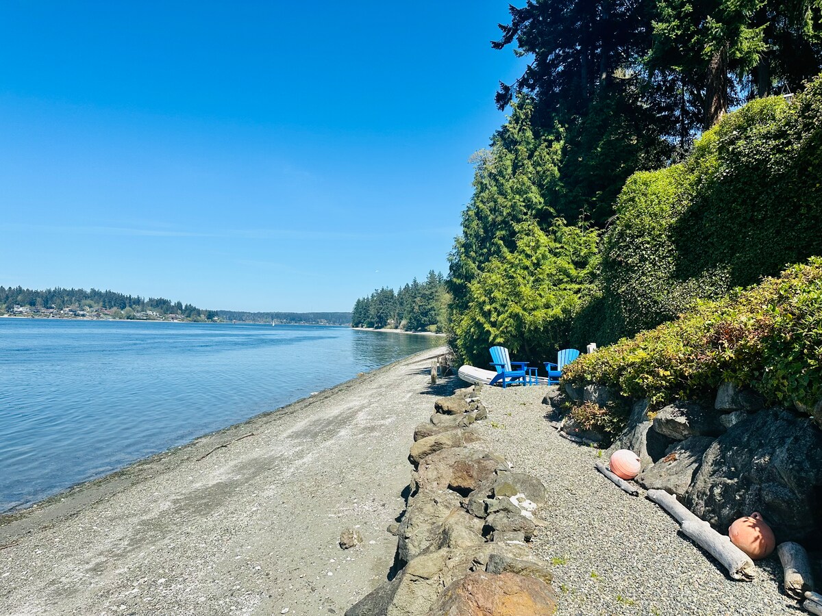 A serene shoreline is visible, characterized by smooth pebbles and sandy areas. Lush greenery frames the scene, while a few blue chairs are positioned for relaxation. In the background, calm waters reflect the blue sky, enhancing the peaceful coastal setting.