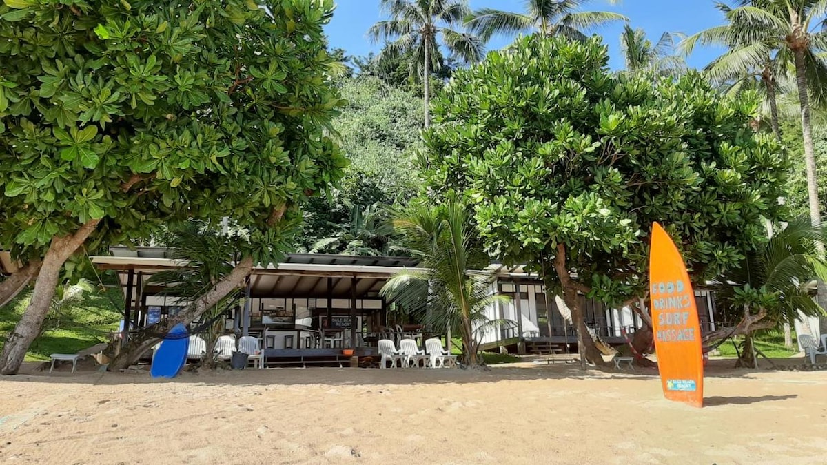 A beachfront bungalow is surrounded by lush greenery and palm trees, offering direct access to the sand. Outdoor seating is arranged on a terrace, while brightly colored surfboards add a playful touch to the entrance. The structure features a large overhang for shade.
