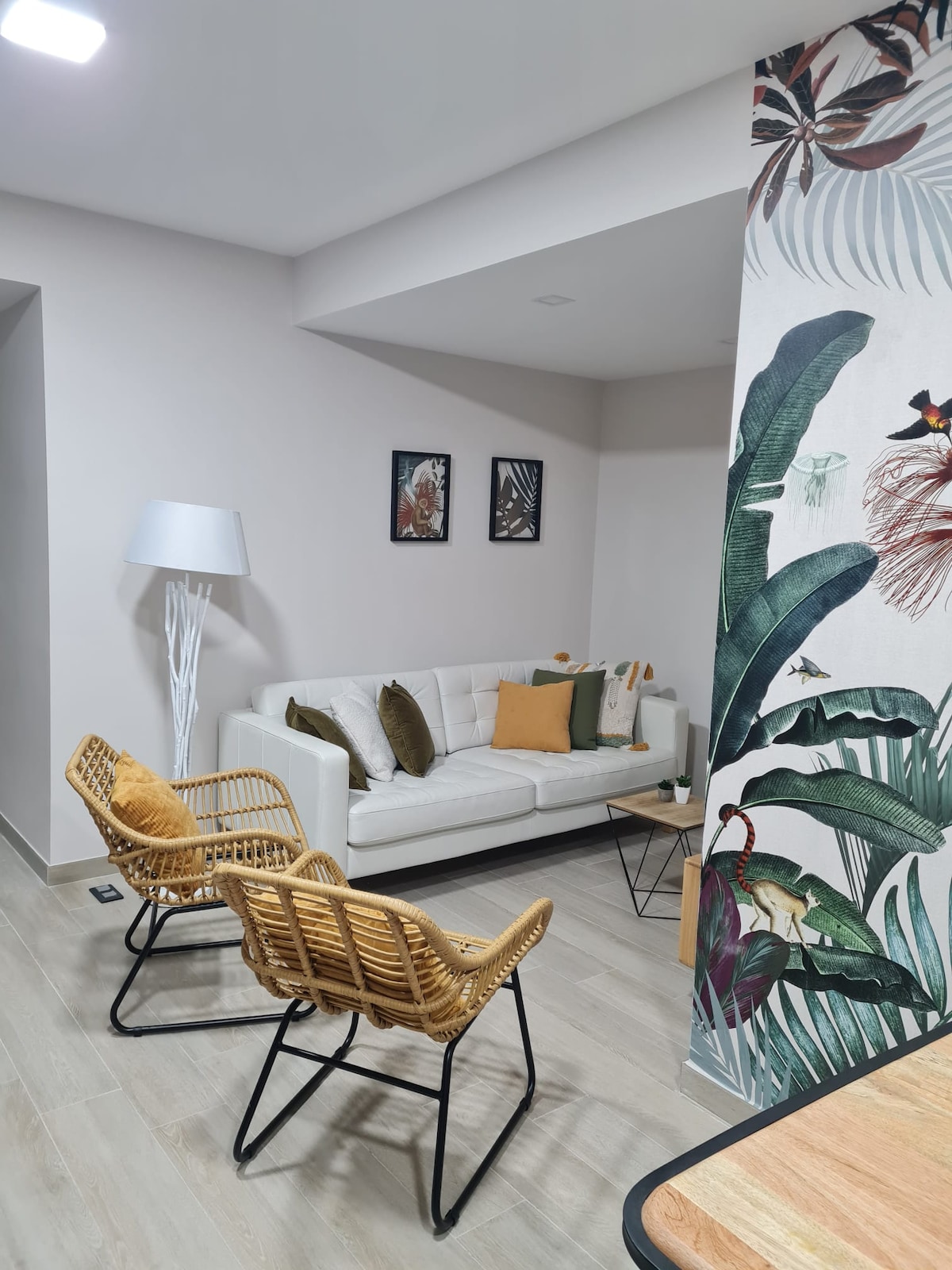 A cozy living area is presented with a white sofa adorned with decorative pillows. Two rattan chairs complement a wooden side table. A tall standing lamp provides soft lighting, while a vibrant wall featuring tropical foliage and bird motifs adds a colorful accent.