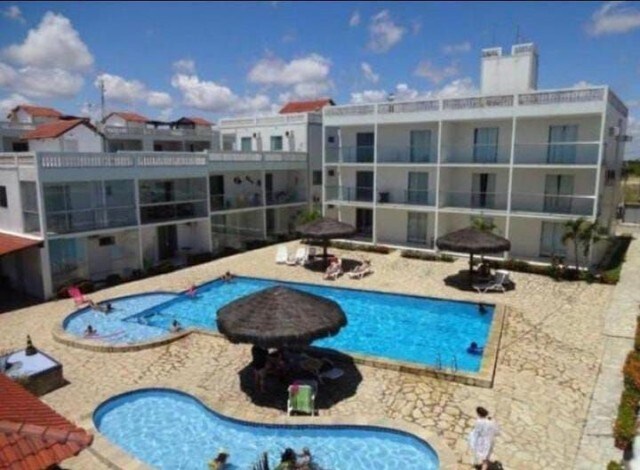 A spacious outdoor pool area is surrounded by loungers and shaded by thatched umbrellas. The pool features a distinct shape and is complemented by a clear patio area made of textured stone. Nearby, two buildings provide views from balconies overlooking the pool.