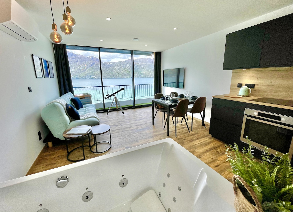 The spacious living area features floor-to-ceiling windows showcasing views of the lake and mountains. A comfortable light blue sofa is positioned near a coffee table. A dining table set for four is visible beside a modern kitchen, with a relaxing hot tub in the foreground.