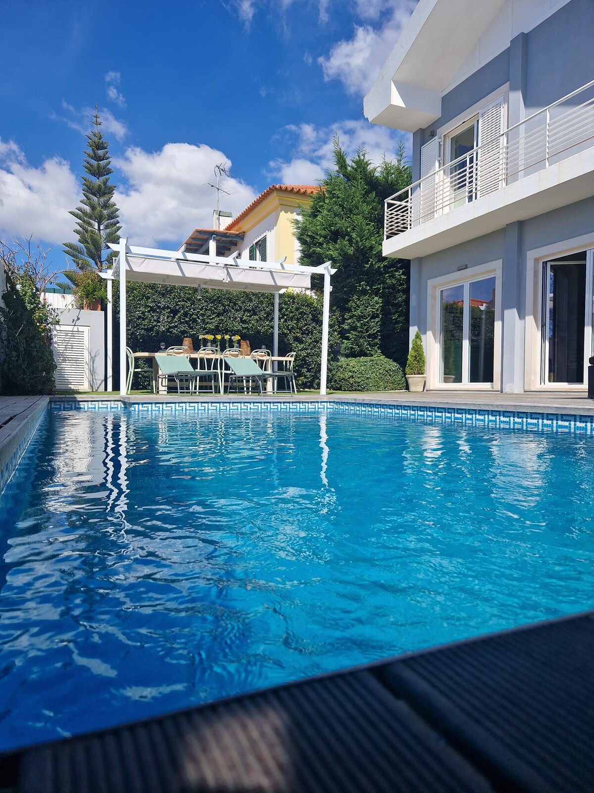 A clear blue pool reflects sunlight under a bright sky. Adjacent, a covered dining area with a large table and several chairs is surrounded by lush greenery, offering a private outdoor setting. The house's exterior showcases modern architectural lines and large windows.