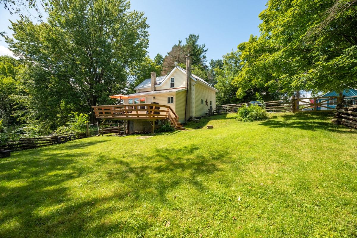 A spacious lawn surrounds the cottage, which is painted in soft yellow and features a deck with a wooden railing. Lush trees provide shade, and a small fenced area is visible, enhancing the privacy of the outdoor space.