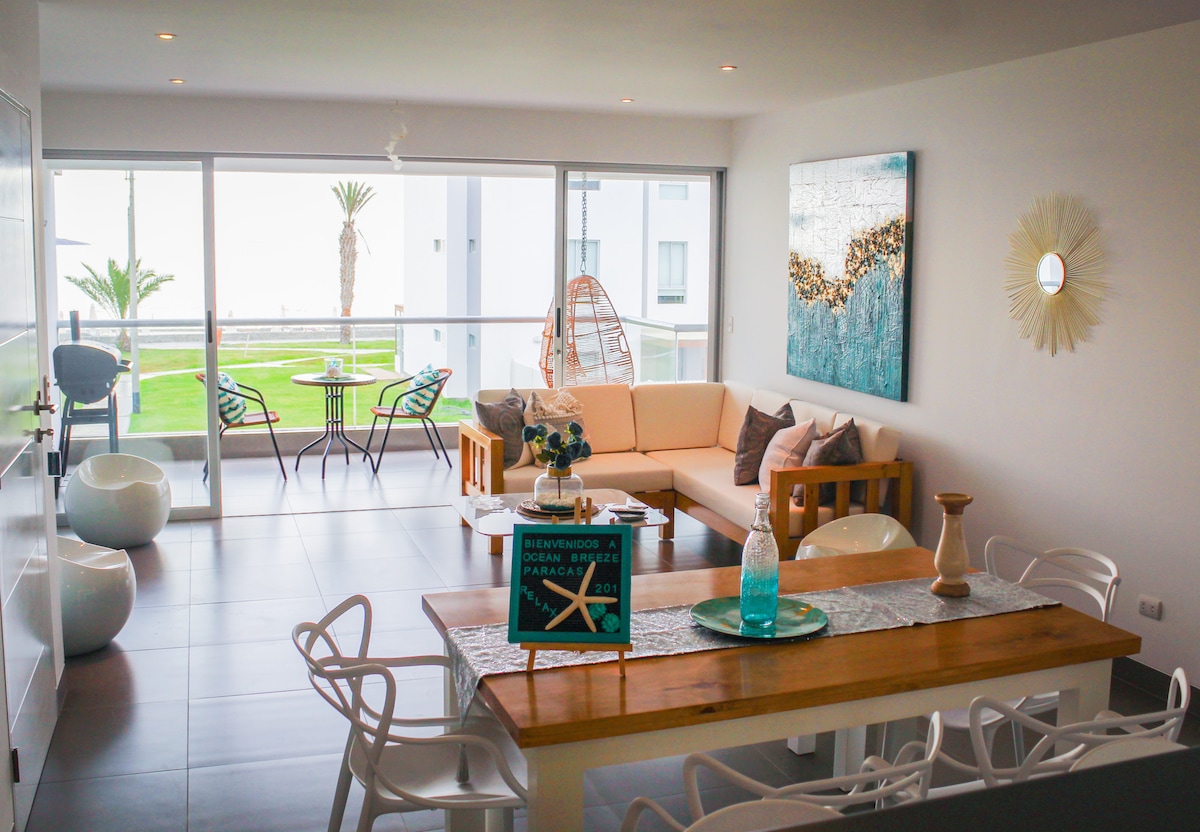 The living area features a light-colored sofa and wooden accents, complemented by a dining table with white chairs. Large glass doors allow for ample natural light and provide views of palm trees and the ocean. A decorative wall piece adds a touch of color.
