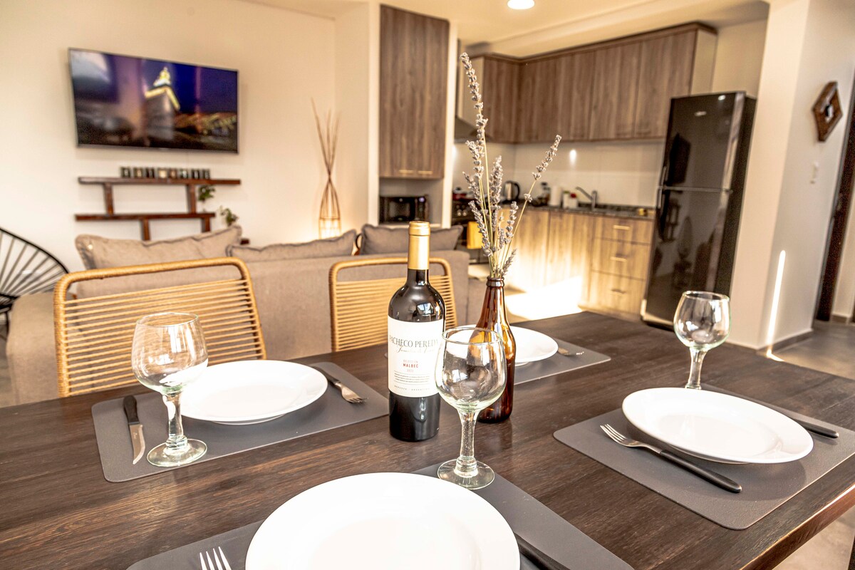 A dining area is set for a meal, featuring a dark wooden table with plates and wine glasses arranged neatly. In the background, a modern kitchen is visible, complemented by a living space with neutral-colored furniture and a large TV on the wall.