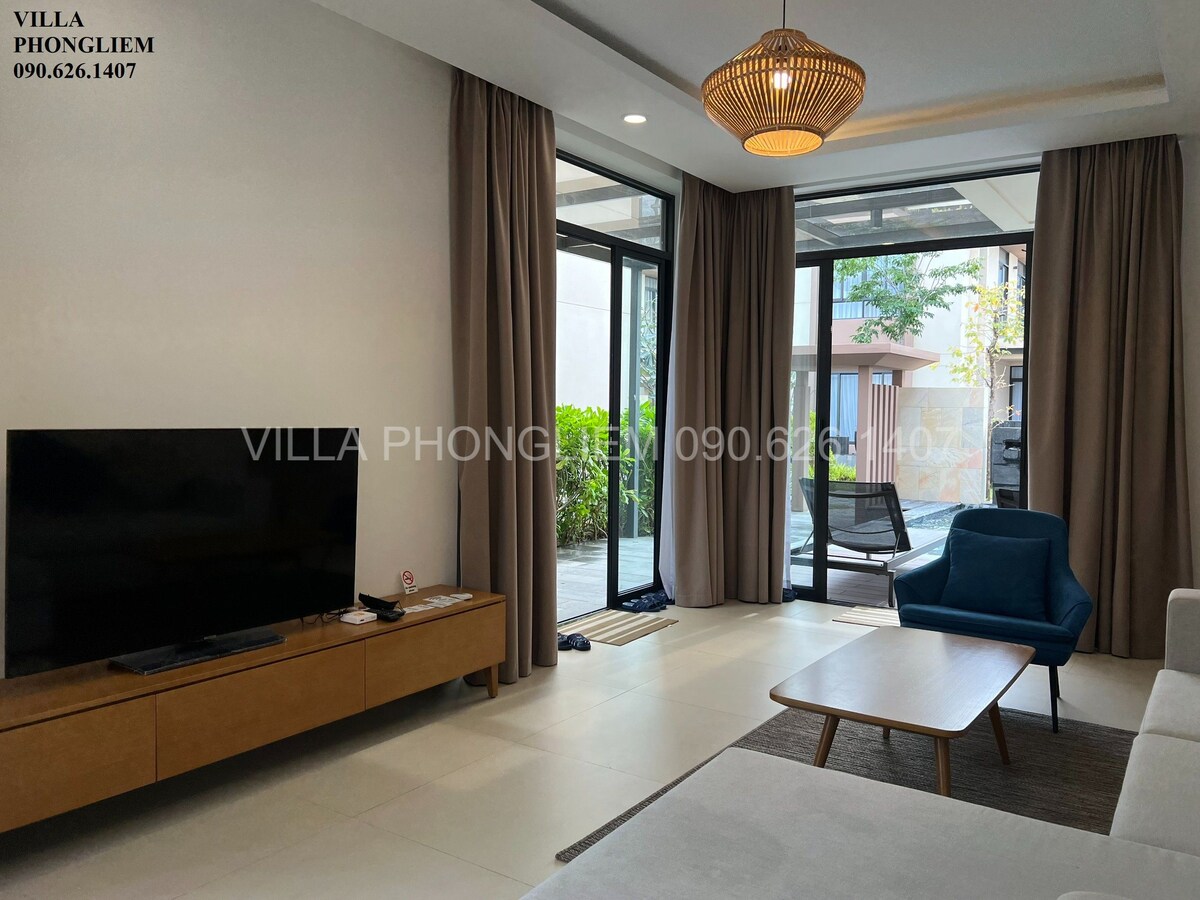 The living area features large glass doors that open to a garden view, allowing natural light to fill the space. A comfortable blue chair is positioned beside a low wooden table, while a large flat-screen TV is mounted on a wooden media console. Soft curtains frame the windows.