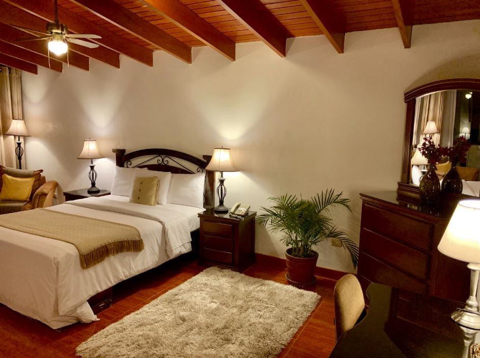 A well-appointed room features a queen bed with a neutral-colored blanket. Soft lighting from two bedside lamps creates a calming ambiance. A wooden dresser and mirror complement the space, while a small desk sits against the wall. A potted plant adds a touch of greenery.