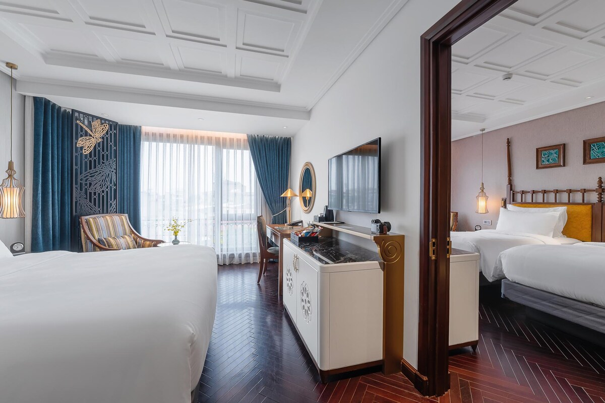 A well-appointed guestroom features a spacious bed and a separate area with a cozy armchair by the window. Natural light enters through sheer curtains, illuminating the elegant décor, which includes a work desk and a flat-screen television against a backdrop of rich wood and soft textiles.