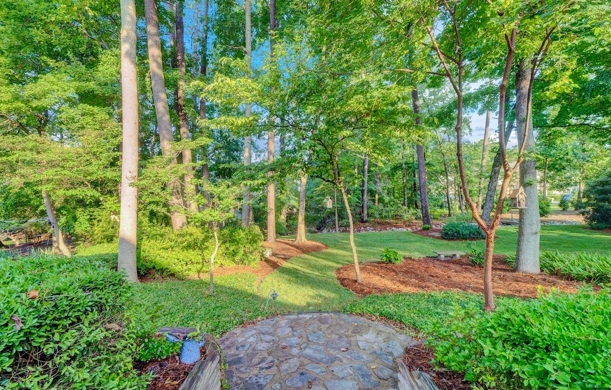 A natural pathway made of stone leads through a partially wooded yard, surrounded by lush greenery. Tall trees offer shade, while manicured bushes line the edges. Sunlight filters through the leaves, creating a serene atmosphere in the inviting outdoor space.