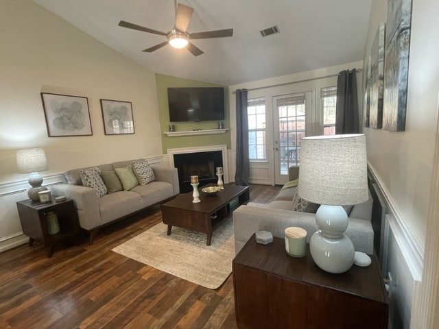 A living space is presented with a light and airy feel, featuring two sofas arranged around a central table. A flat-screen TV is mounted above a fireplace. Natural light filters in through patio doors leading to a private outdoor area, framed by curtains.