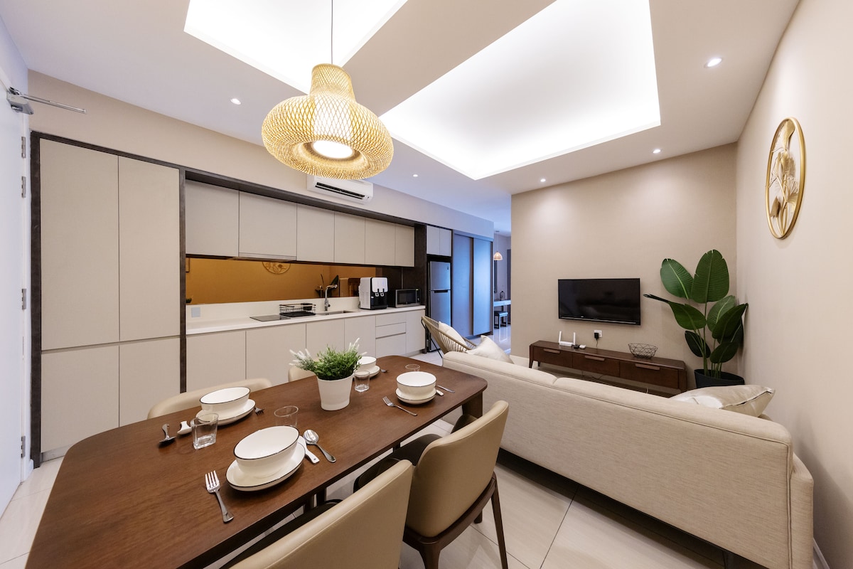 A contemporary open-plan living and dining area is featured, illuminated by soft lighting. A wooden dining table is set with dinnerware, while a comfortable sofa is positioned across from a mounted television. A potted plant adds a touch of greenery to the neutral-toned decor.
