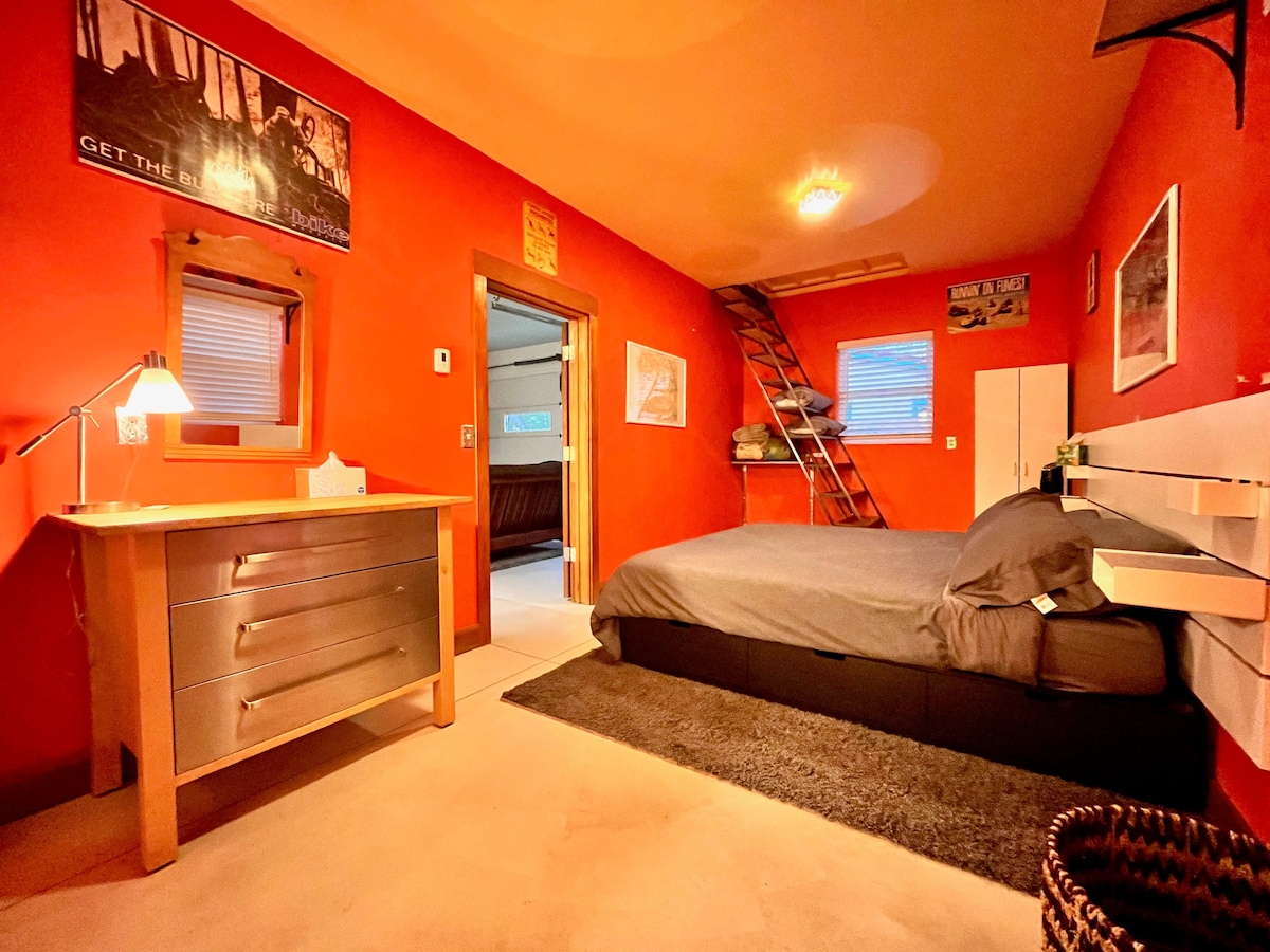 A vibrant bedroom features warm red walls and a spacious King Bed with simple bedding. A wooden dresser is positioned beside a desk lamp, while a ladder leads to a lofted sleeping area. Natural light streams through a window, enhancing the room's inviting ambiance.