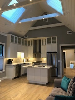 A modern kitchen is featured with sleek white cabinetry and stainless steel appliances. Skylights allow natural light to fill the space, highlighting the open layout. A central island offers additional counter space, complemented by a cozy seating area with a grey sofa and teal accent pillows.