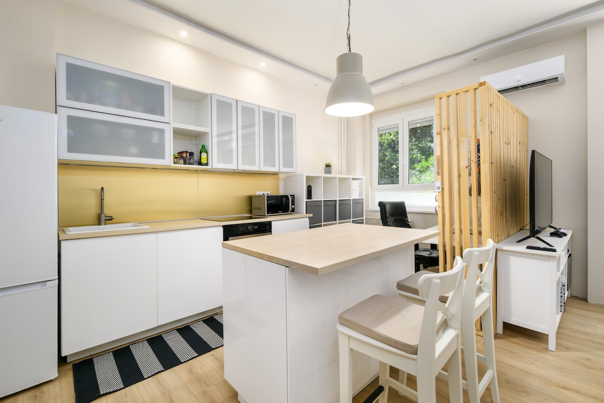 The kitchen area features sleek white cabinetry with a light wood countertop and an island with two stools. A microwave and refrigerator are present, alongside a black chair and a partition separating the workspace. Natural light filters in through the window overlooking the green courtyard.