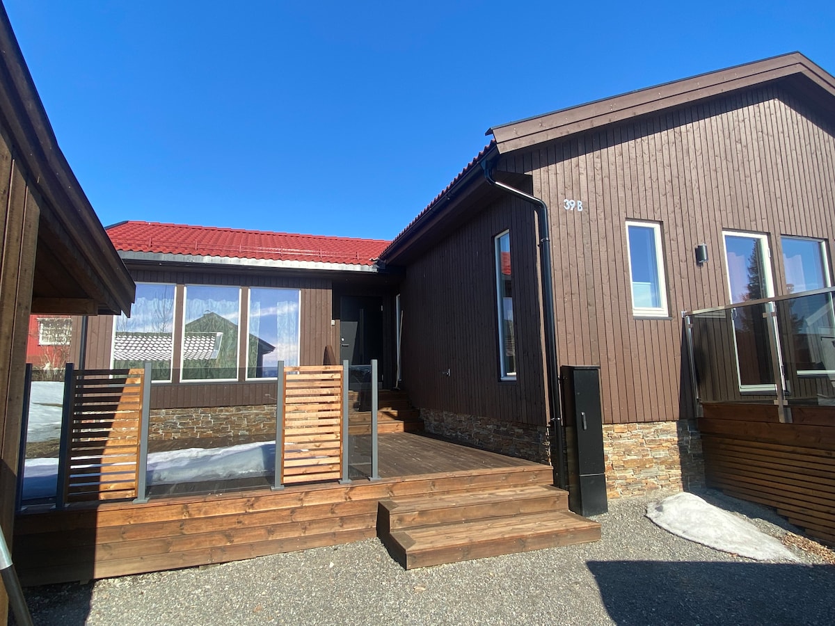 A modern wooden house features a large deck with wooden railings and steps leading to the entrance. Expansive windows allow natural light to fill the interior, while a gravel area surrounds the home. The clear blue sky creates a bright atmosphere.
