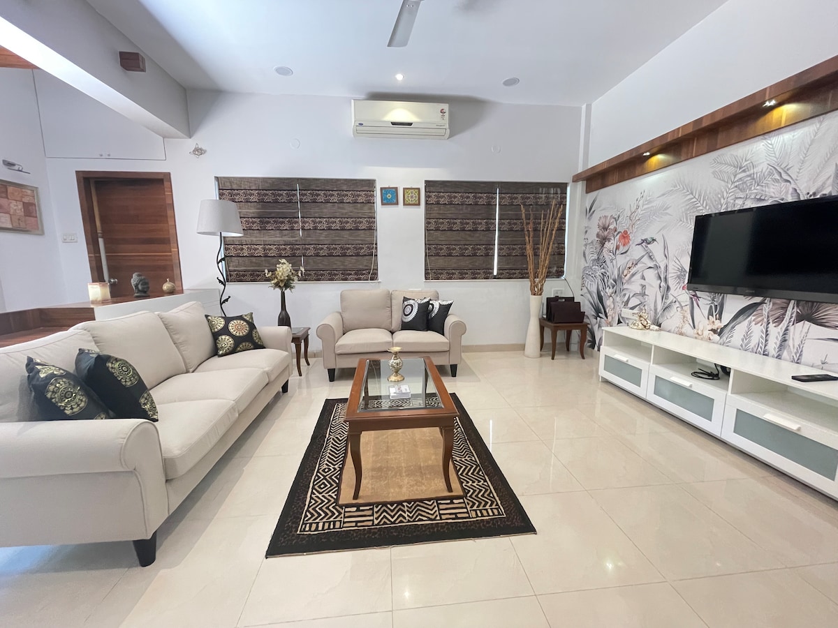 A spacious living area features a light-colored sofa, a coffee table with decorative items, and a television mounted on the wall. Natural light enters through windows adorned with wooden blinds, while tasteful wall art and a patterned rug enhance the ambiance.