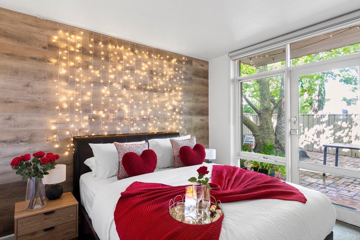 A cozy bedroom features a spacious bed adorned with decorative pillows and a vibrant red throw. Soft, warm fairy lights illuminate an accent wall, enhancing the romantic atmosphere. Large windows provide views of the garden, allowing natural light to fill the room.