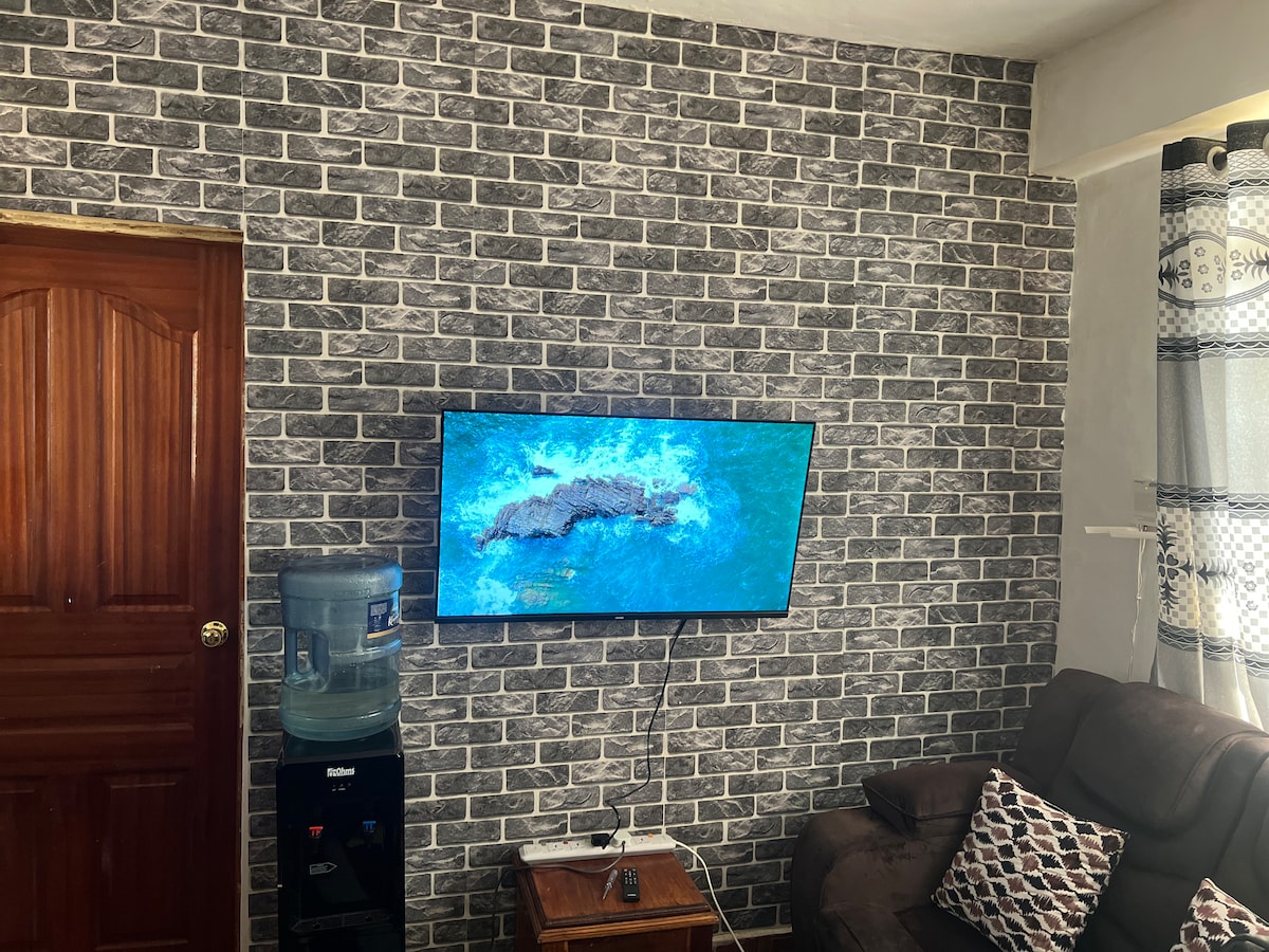 A modern living area features a grey brick wall adorned with a large television, displaying a vibrant blue image. A water dispenser stands beside a wooden side table, while a comfortable brown sofa is complemented by a decorative cushion.