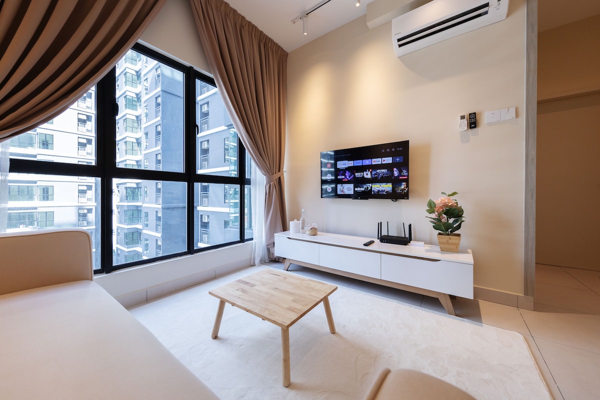 The living room features large windows allowing natural light to fill the space. A minimalistic coffee table is positioned in front of a comfortable sofa. A Smart TV mounted on the wall showcases a selection of entertainment options, alongside a decorative plant atop a media console.