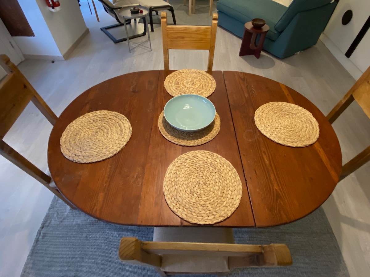 An oval wooden dining table is centered in the space, surrounded by four wooden chairs. The table is set with a large blue bowl and five round woven placemats arranged evenly. A neutral-toned rug is placed underneath, enhancing the dining area.