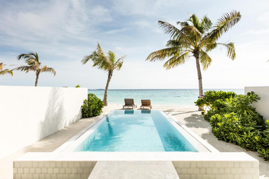 A tranquil private pool is surrounded by lush greenery and gently swaying palm trees, leading to a sandy beach and the ocean beyond. Comfortable loungers are positioned by the pool, inviting relaxation with a peaceful coastal view.