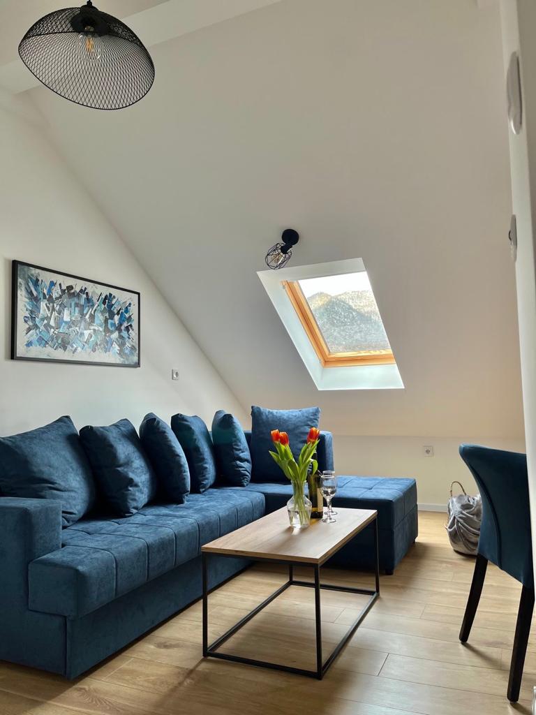 A bright living space features a blue sectional sofa against a slanted wall, complemented by a wooden coffee table. A vase of fresh tulips is displayed centrally. A skylight above allows natural light to illuminate the area, enhancing the airy atmosphere.