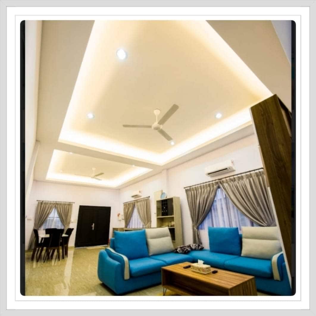 The living area features a spacious layout with modern furnishings. A large blue sectional sofa is positioned in front of a wooden coffee table. Soft lighting is provided by recessed ceiling lights, and ceiling fans are seen above. Curtains frame the windows, enhancing the welcoming atmosphere.
