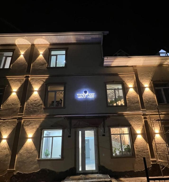 The exterior of Bay Ali is illuminated at night, showcasing the elegant facade with bright lights highlighting the architectural features. Large windows reflect the soft glow, and the entrance is framed by a welcoming doorway, inviting guests to explore the accommodation.
