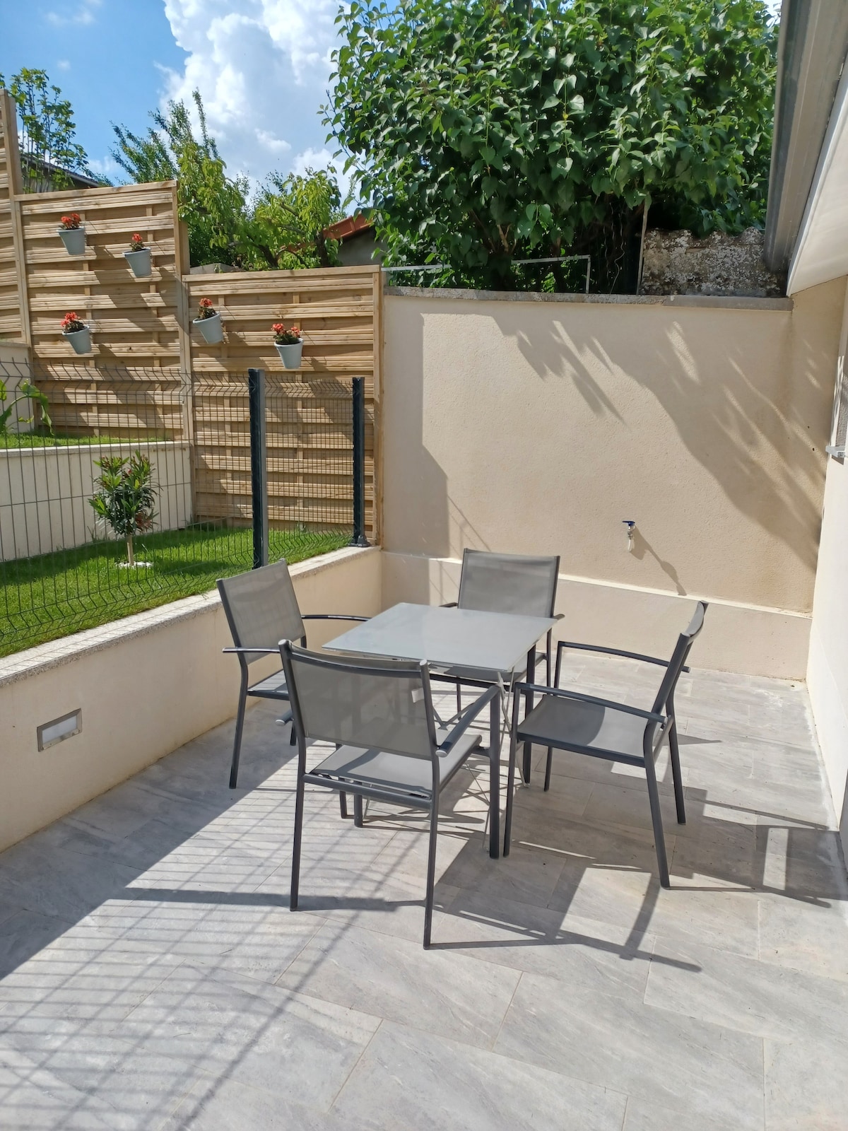 A patio area is seen, featuring a gray dining table surrounded by four matching chairs. The space is enclosed by a low wall and a fence with greenery visible in the background. Natural light illuminates the area, enhancing the inviting setting.