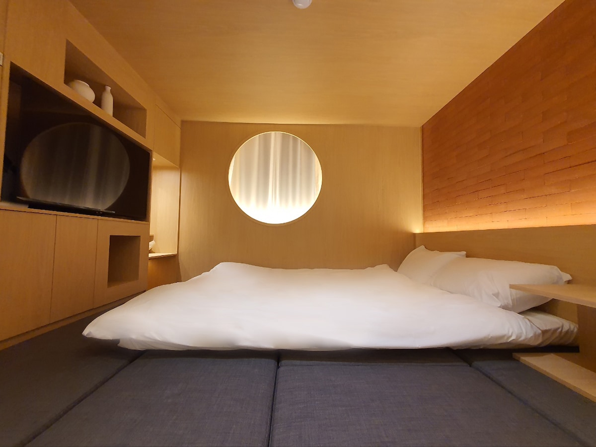 A minimalist sleeping area is presented, featuring a spacious mattress atop soft, dark bedding. The warm wood tones complement a round window that filters soft light into the room. Shelving provides a place for decor, and a flat-screen TV is positioned on the side.