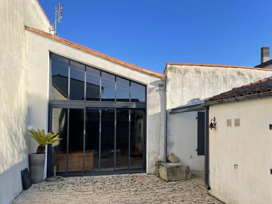 A spacious exterior view of the property is presented, featuring a large glass wall that allows natural light to illuminate the interior. A paved courtyard is visible, bordered by white walls and a terracotta roof, enhancing the peaceful ambiance.