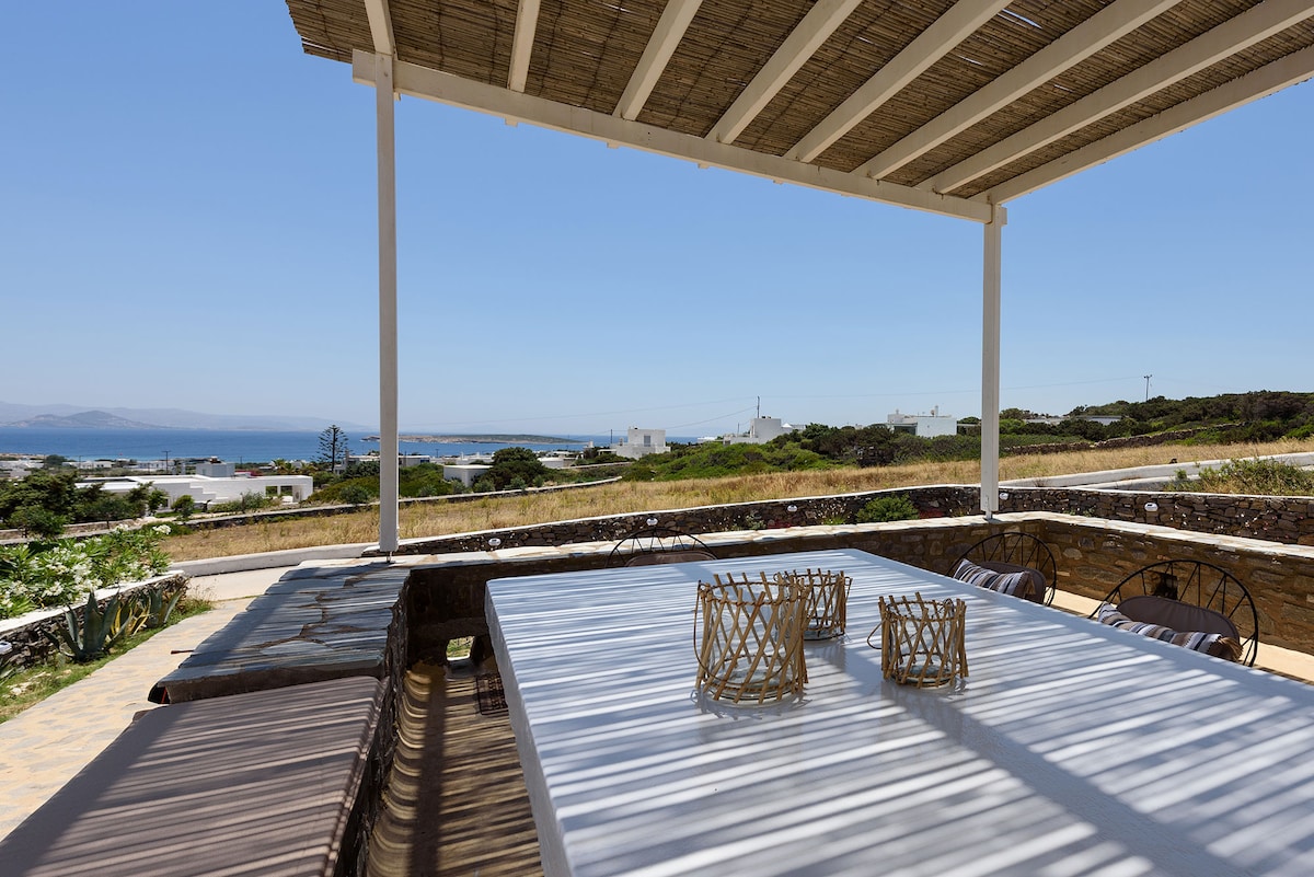 An outdoor dining area is showcased under a slatted pergola, featuring a long white table surrounded by round chairs. Natural light creates a dappled effect on the table surface, while a view of the sea and distant landscape is visible in the background.
