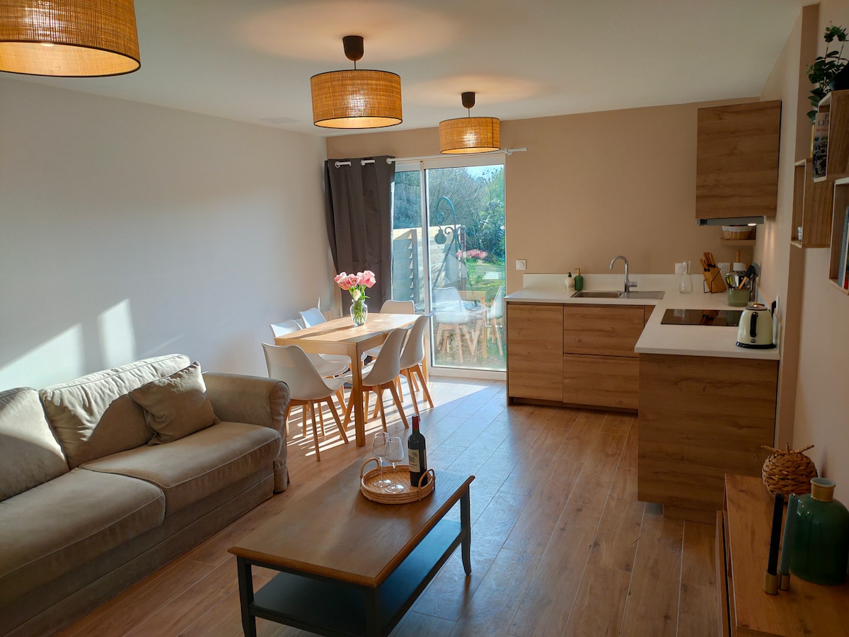 A bright and spacious living area features a comfortable sofa beside a wooden coffee table. A dining table with white chairs is set near a glass door, allowing natural light to illuminate the space. Modern cabinetry is visible in the kitchen area.