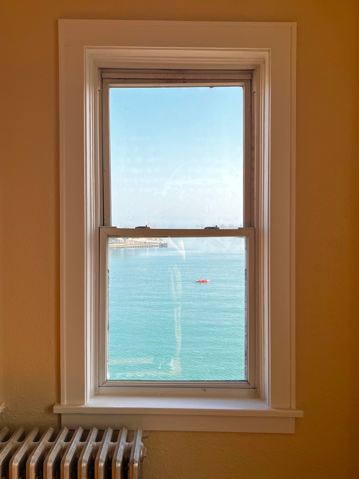 A window framed by white trim offers a view of Lake Michigan. The calm waters reflect the sky, with a small boat visible in the distance. The walls are painted a warm yellow, complementing the natural light entering the space.