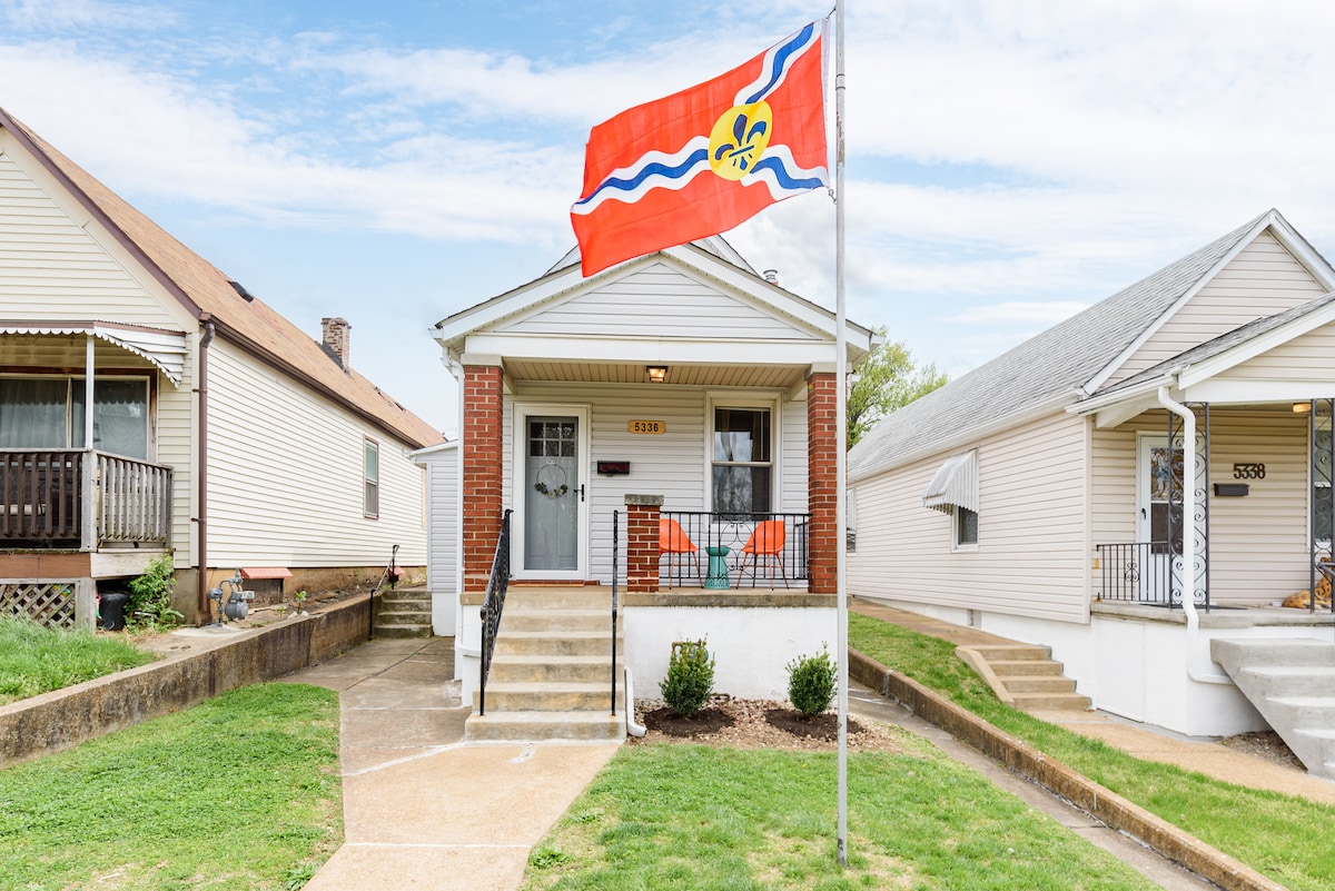 A charming bungalow is showcased with its welcoming front porch, featuring steps leading up to the door. The porch is adorned with orange chairs, and a bright flag is displayed prominently on a pole, complementing the well-maintained lawn and surrounding homes.