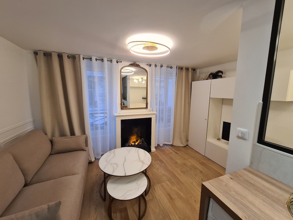 A cozy living area features a simple beige sofa positioned opposite a modern fireplace. A marble coffee table sits in front, complementing the light wood flooring. Sheer curtains allow natural light to filter in, enhancing the room's warm atmosphere.
