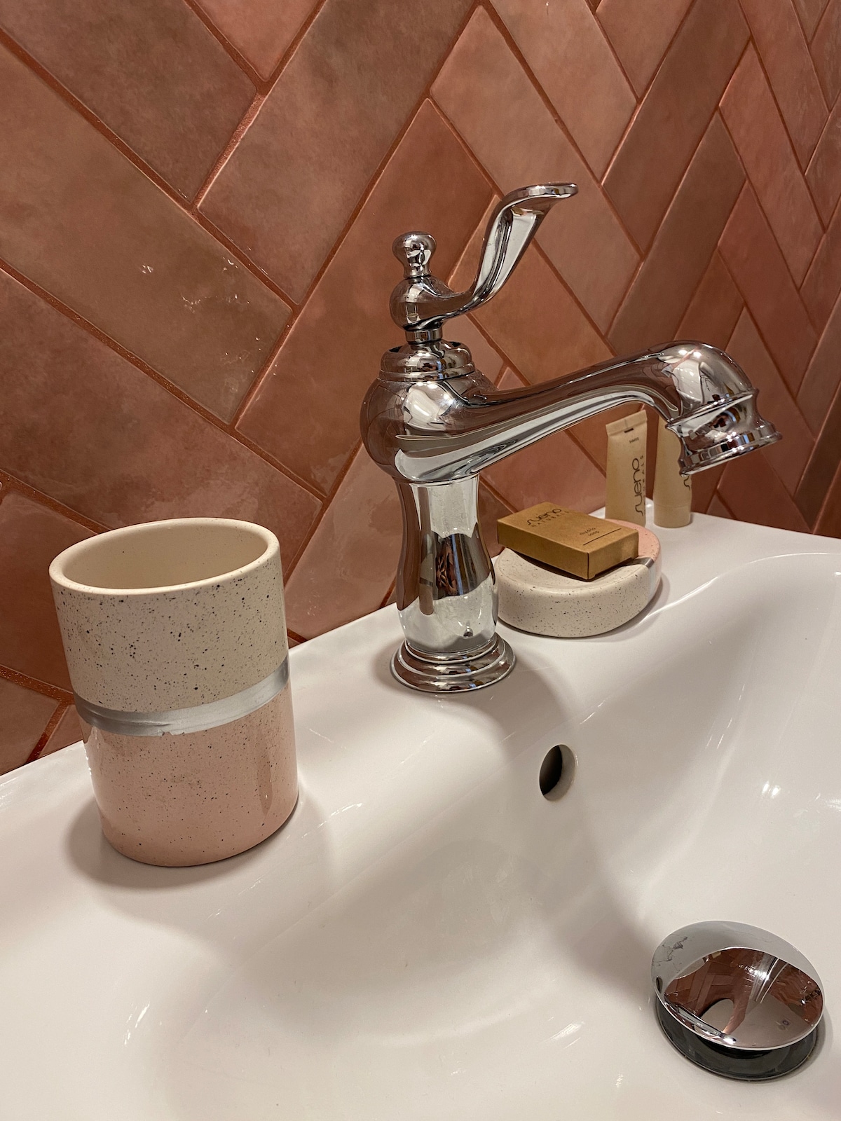 A modern bathroom sink is featured, showcasing a chrome faucet with a vintage design. Beside the faucet, a two-tone ceramic cup and a small decorative dish hold toiletries. The backsplash consists of pink herringbone tiles that add texture to the space.