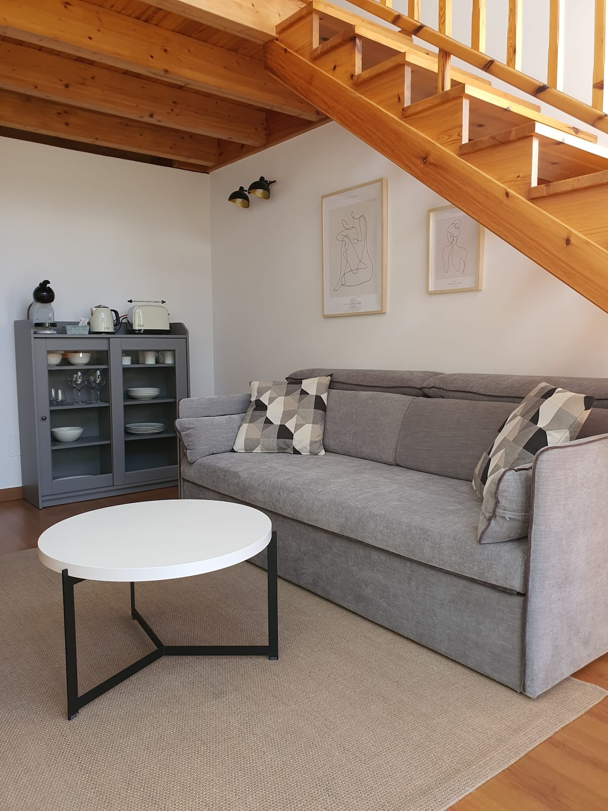 A living area is depicted with a gray sofa adorned with patterned cushions, positioned adjacent to a wooden staircase. A round white coffee table sits in front of the sofa. A gray cabinet is visible in the background, containing dishware and appliances.