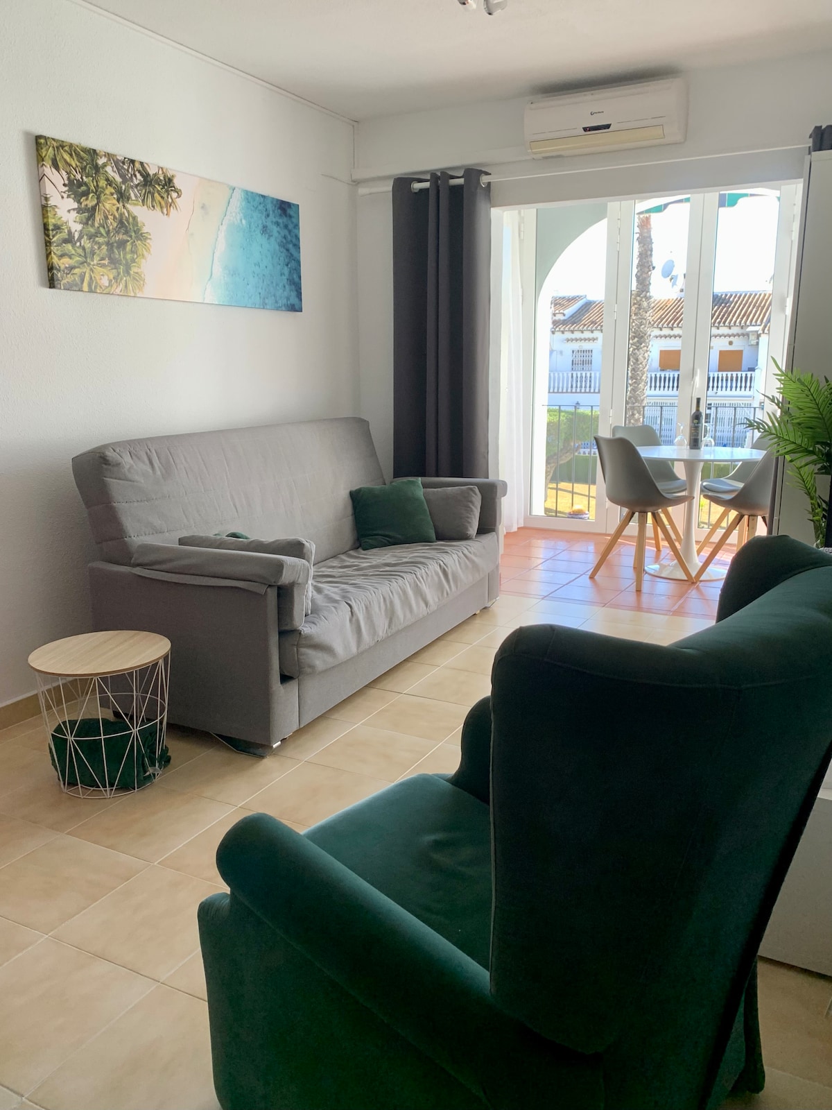 A living area features a gray fold-out sofa and a green armchair, positioned near a set of glass doors that lead to a small balcony. A round table with two chairs is visible, alongside decorative plants and light-colored tiled flooring.