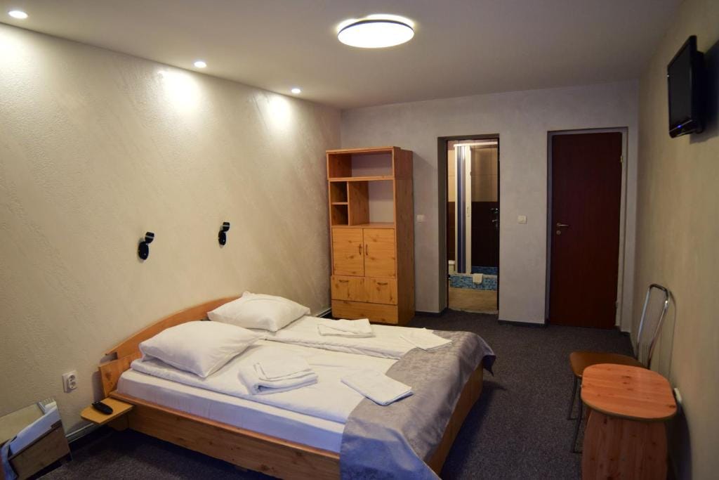 A cozy sleeping area is featured with a spacious bed, neatly arranged linens, and two reading lights mounted above. A wooden shelving unit and a small table are included, while an adjoining door leads to a bathroom. Neutral walls create a calm environment.