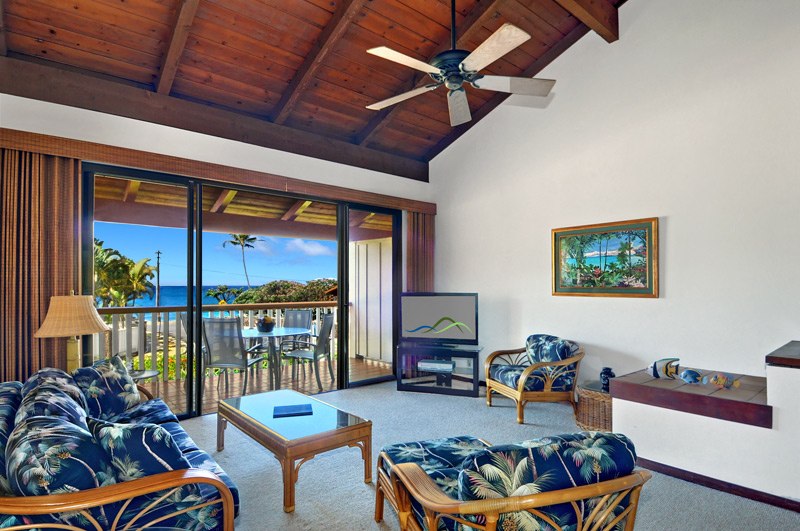 A welcoming living area features a high wooden ceiling and large glass doors that open to a balcony with ocean views. Tropical-patterned furniture is arranged around a coffee table. A flat-screen TV and decorative artwork complete the comfortable space.