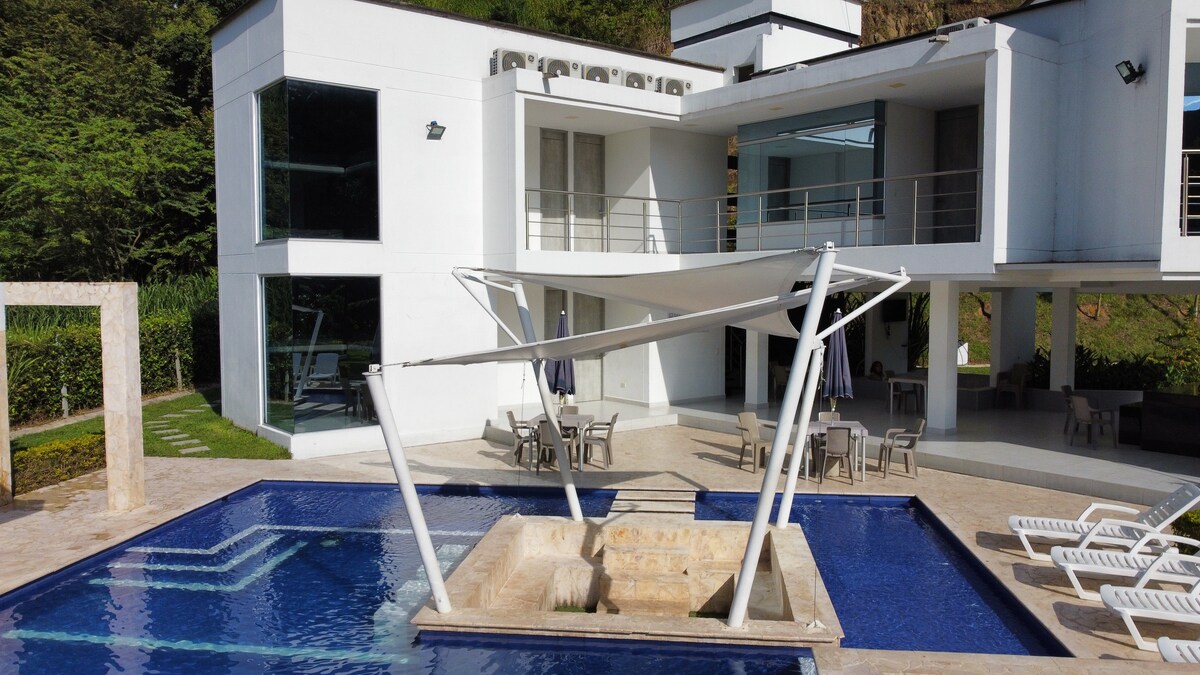 A modern minimalist structure is shown, featuring expansive glass windows and an open layout. A spacious pool with a built-in jacuzzi is visible in the foreground, along with a shaded seating area and lounge chairs arranged around the pool's edge.