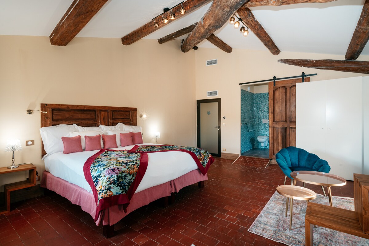 A spacious bedroom features a large bed adorned with a decorative blanket. Exposed wooden beams add character to the ceiling, while a stylish blue chair complements wooden furniture. A private bathroom with a glass partition is visible in the background, highlighted by blue tiles.
