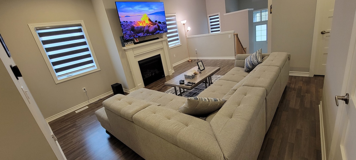 A spacious living area is showcased with a large sectional sofa and a coffee table in front. A fireplace adds a cozy element, while a large television mounted above displays a scenic image. Large windows with striped blinds allow natural light to fill the room.
